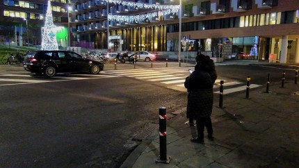 Person standing at the side of a street at night looking at their mobile device. Pedestrian crossing and a car in the background.  