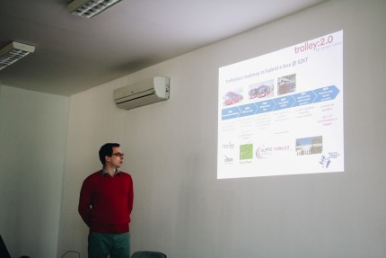 A man is giving a powerpoint presentation that is being projected onto a white wall. A small AC can be seen to the left of the projection. 