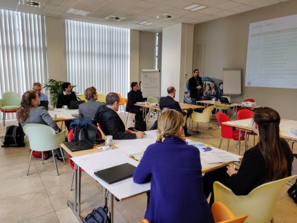 People sitting in groups of 4 in a well lit room at desks with their laptops and notebooks open, listening to a man giving a presentation 