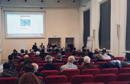 Audience members sitting in rows of theatre seats looking at a presentation from a projector with four people sitting in a panel on a slightly raised stage platform 