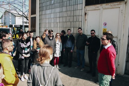 People standing in a circle outside an industrial building listening to man speaking to the whole group 