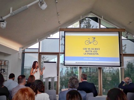 A woman giving a speech to a croud with a projection screen showing a bicycle and an Italian slogan 