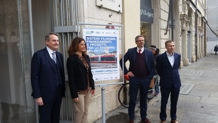 Four government officials standing in front of poster announcing national trolleybus day in Parma. Woman trying to unlock her bicycle directly behind them. 
